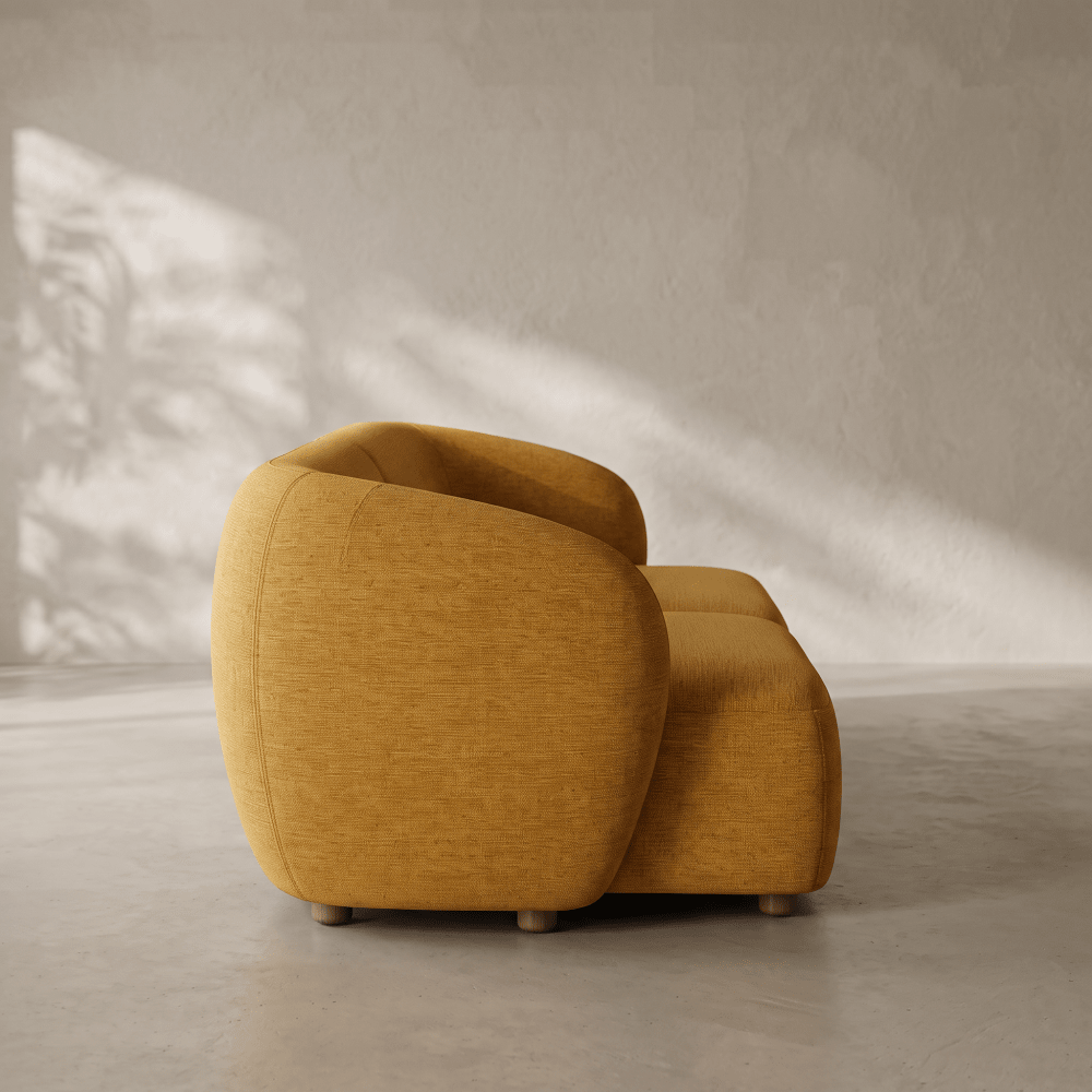 A modern curved armchair upholstered in textured mustard yellow fabric, shown in a minimalist room with concrete floors and textured walls under soft diagonal sunlight.