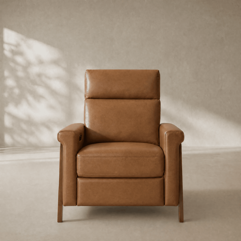 A tan leather recliner armchair with a wooden frame, positioned centrally against a textured beige wall with soft leaf shadows cast across it.