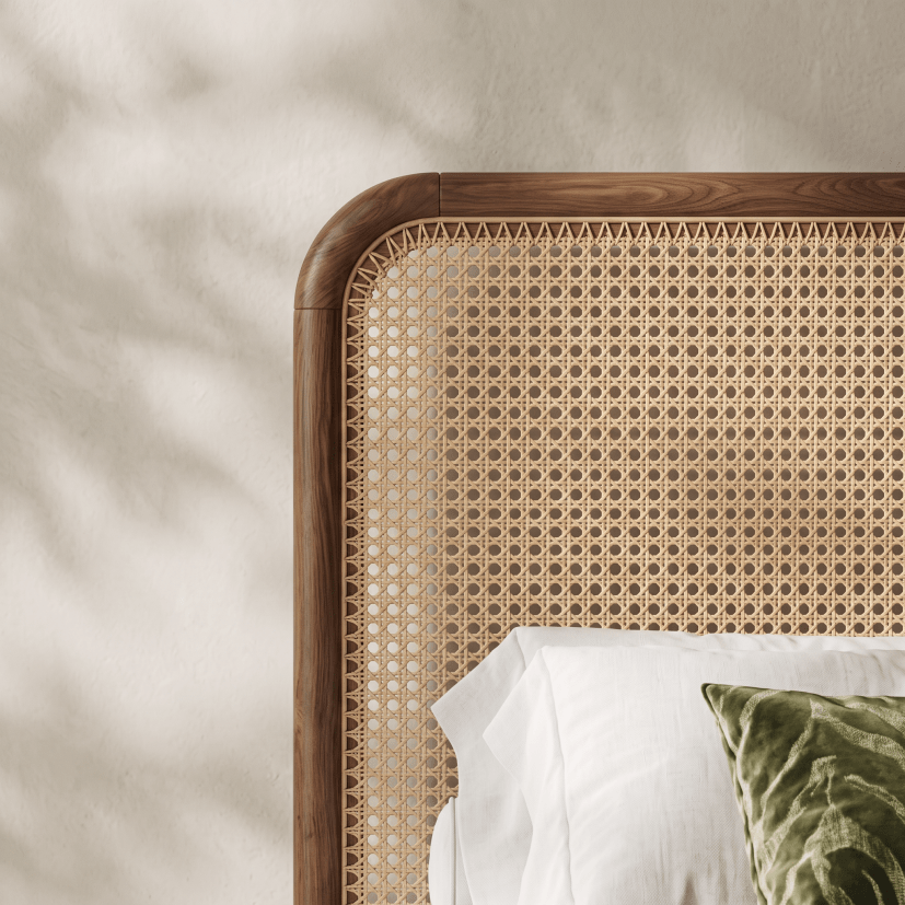 Close-up of a wooden bed headboard with a woven cane or rattan panel, styled with white bedding and a green textured pillow against a warm beige wall with soft leaf shadows.