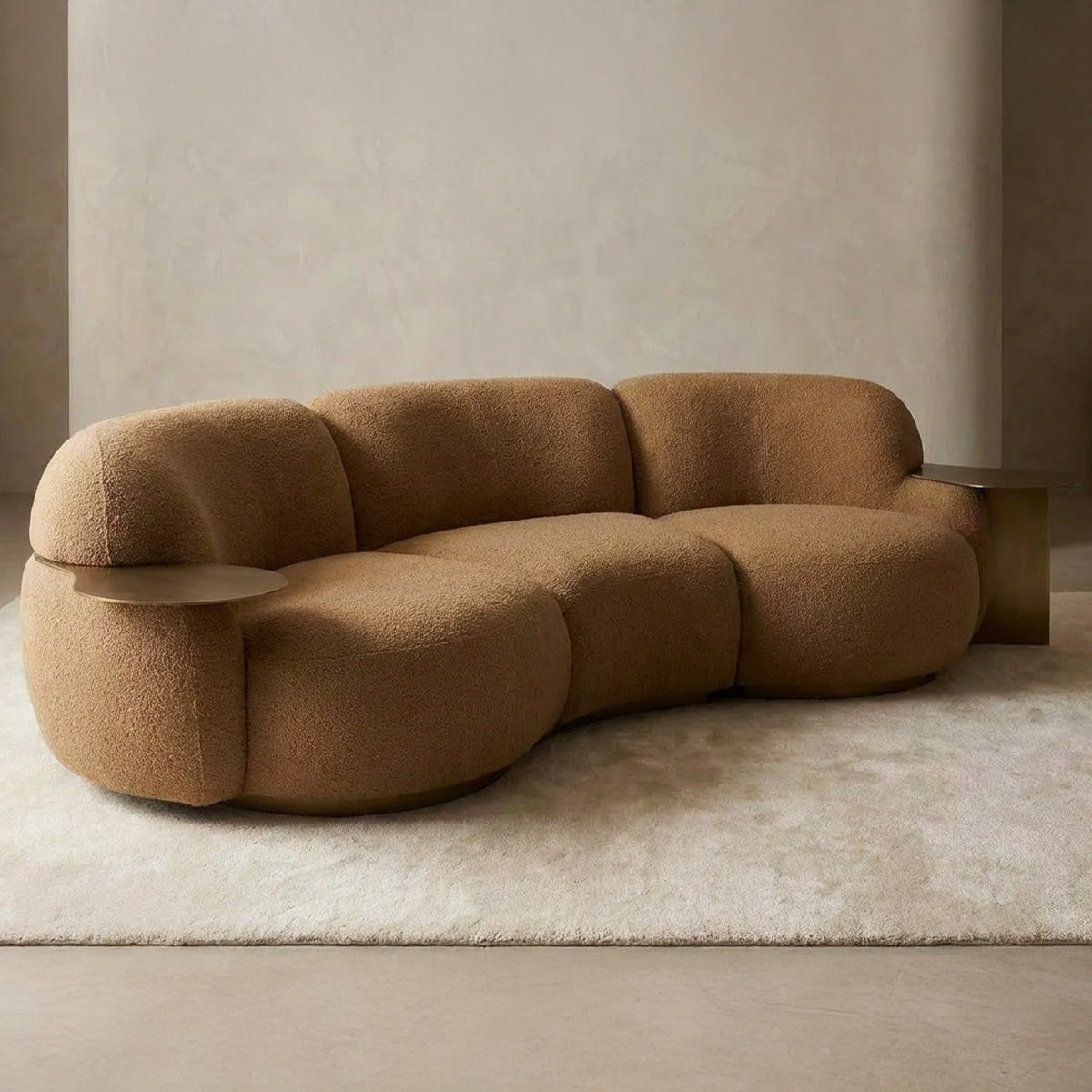 Three-seater curved sofa in caramel bouclé fabric featuring an organic shape, an integrated circular brass side table on the armrest, and a gold metal plinth base.