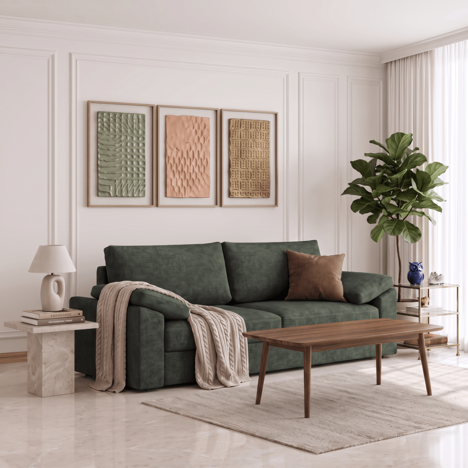 A modern living room featuring a dark green fabric sofa with a brown accent pillow, a beige throw blanket, a wooden coffee table, and three framed textured art prints on a white paneled wall.