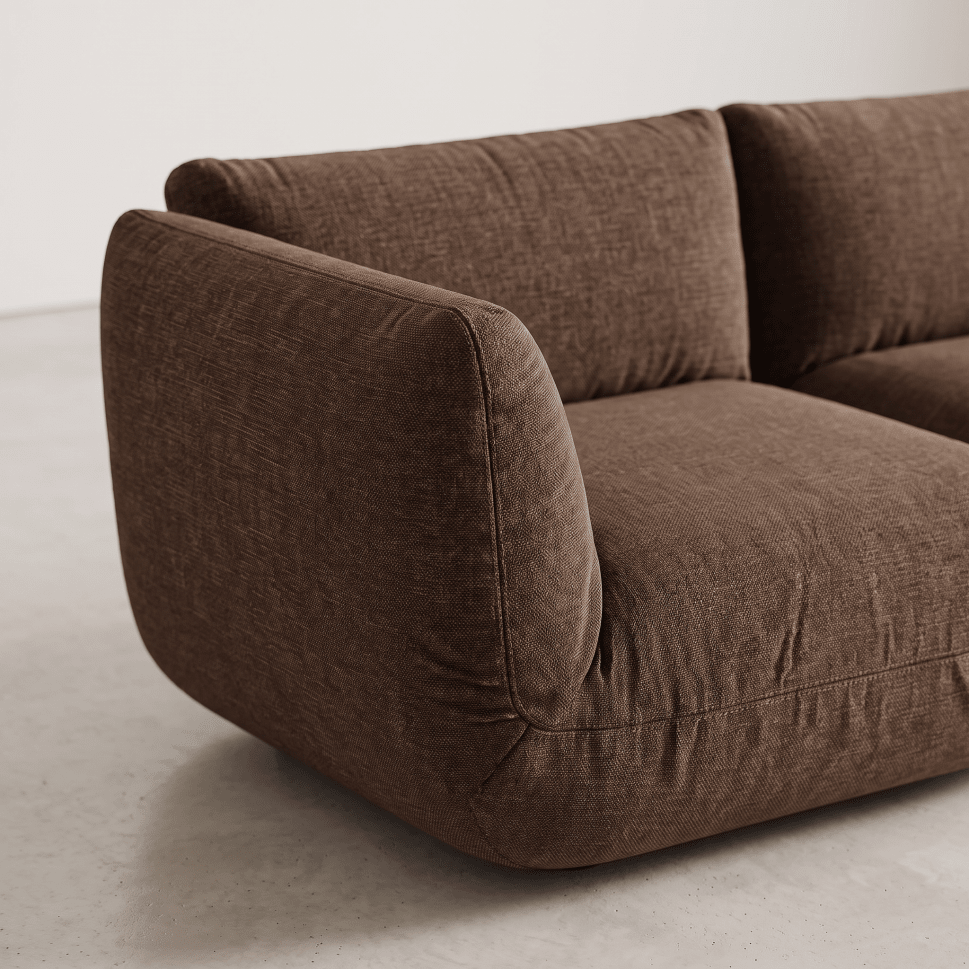 A close-up of a modern brown upholstered sofa corner with rounded arms and a textured fabric, set against a clean light gray wall and floor.