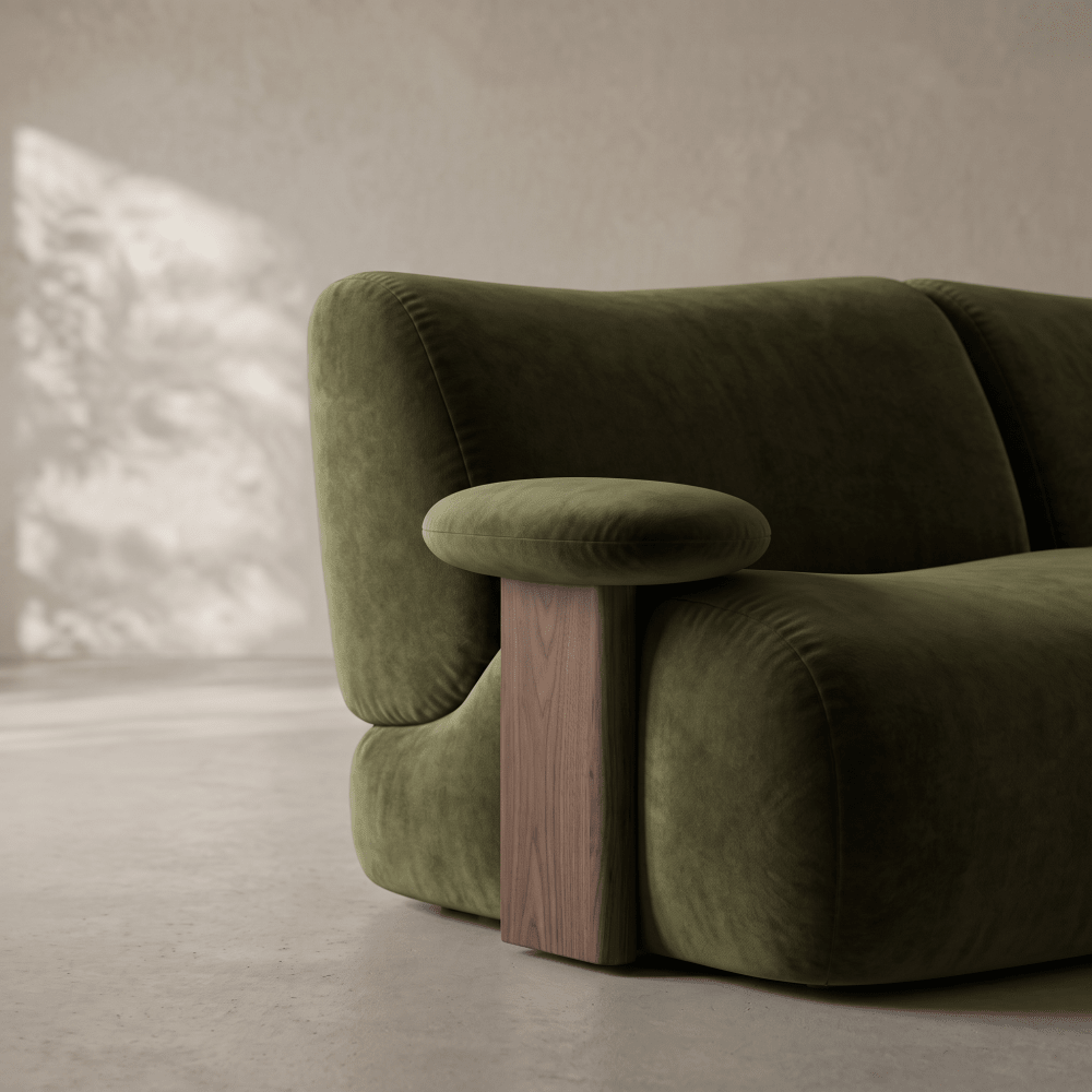 A close-up of a modern green velvet sofa with a chunky wooden leg and rounded armrests, set against a minimalist concrete wall and floor.