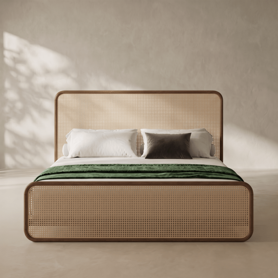 A modern platform bed with a woven cane headboard and footboard, styled with white bedding and a green throw blanket, set against a textured beige wall with soft leaf shadows.