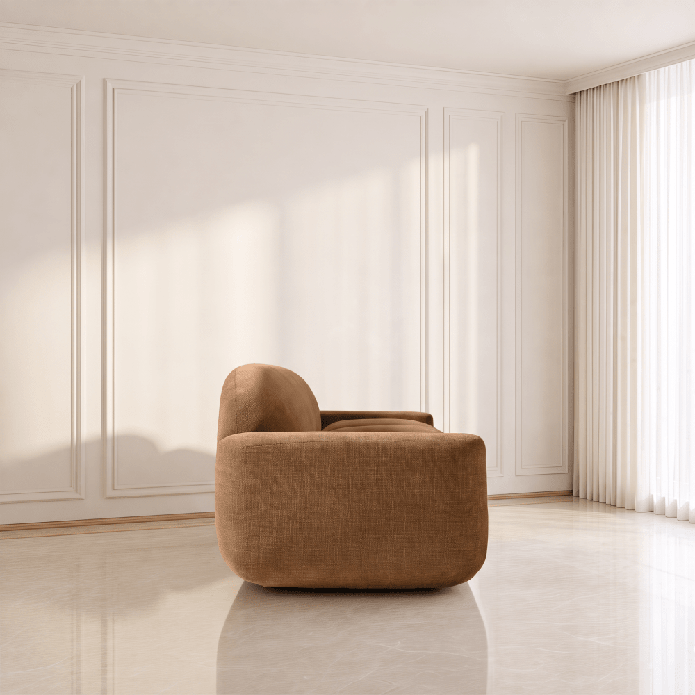 A modern low-profile armchair in a warm brown textured fabric, positioned in a minimalist room with cream paneled walls, a glossy light floor, and sheer curtains.