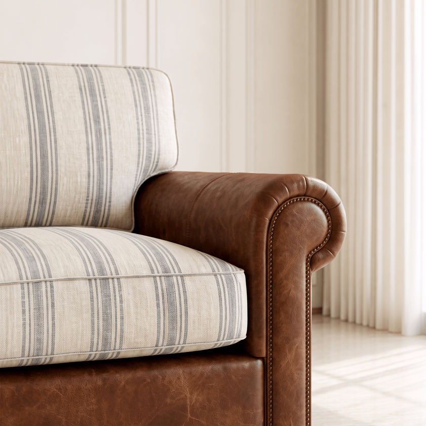 Close-up of a classic armchair corner showing a brown leather rolled armrest with nailhead trim and cream-colored striped fabric cushions.