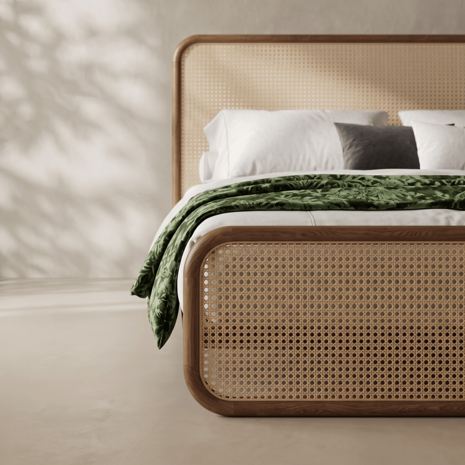 A close-up of a bed frame with a natural cane woven front panel and a matching wooden headboard, styled with white bedding and a green leaf-patterned throw blanket in a softly lit room.